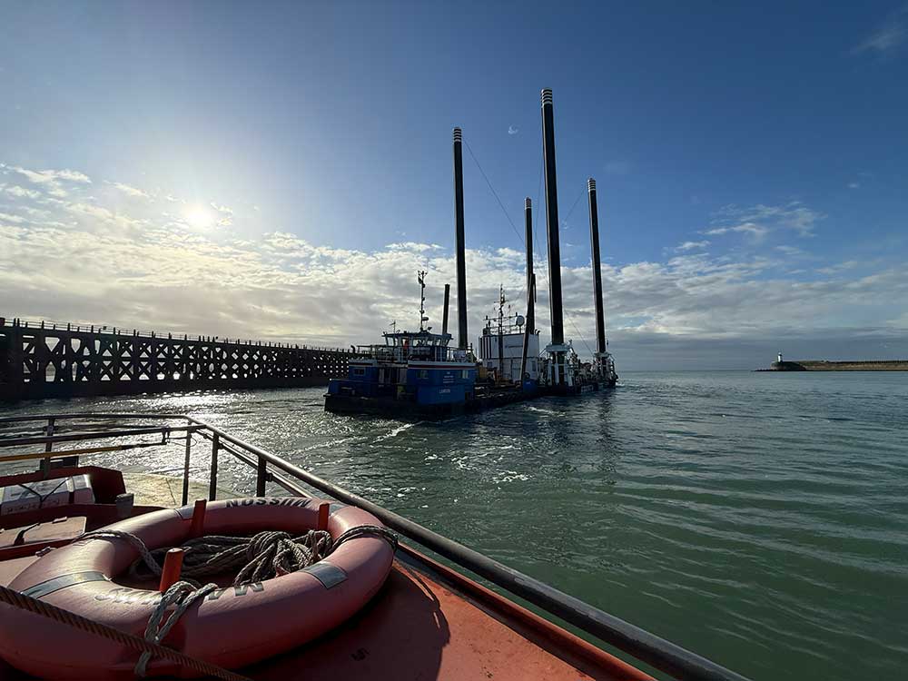 The Octopus platform leaves Newhaven port in the early morning sunlight with the pilot boat in foreground and the lighthouse visible in the distance
